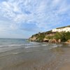 Spiaggia sabbiosa con onde leggere e una casa bianca circondata da vegetazione su una collina.