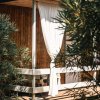 Veranda in legno con tenda bianca tra piante verdi.