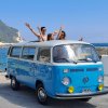 Furgoncino Volkswagen blu vicino al mare. Due persone sporgono dal tetto, godendosi la giornata.