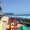 Spiaggia affollata con ombrelloni blu e giochi per bambini sullo sfondo del mare.