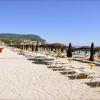 Spiaggia con file di lettini e ombrelloni marroni, vista mare e colline in lontananza.
