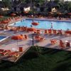 Piscina affollata con sdraio e ombrelloni arancioni, circondata da piccoli edifici e vegetazione.