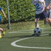 Bambini che giocano a calcio su un campo erboso, vicino a una recinzione.
