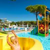 Scivolo giallo con una donna bionda, piscina con giochi d'acqua e resort in background.